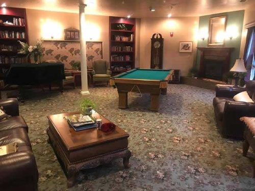 Cozy living room with floral carpet, pool table, leather sofas, and bookshelves. Warm lighting, fireplace, and grand clock create a relaxed, inviting atmosphere.
